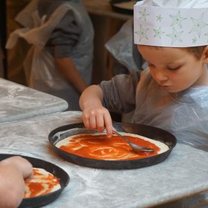 child putting tomato sauce on top of a pizza base