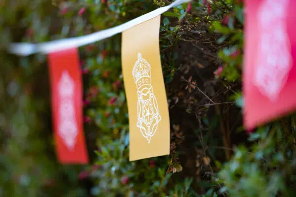 Yellow decorative banner with a white intricate emblem hanging among green bushes, part of a string of festive flags