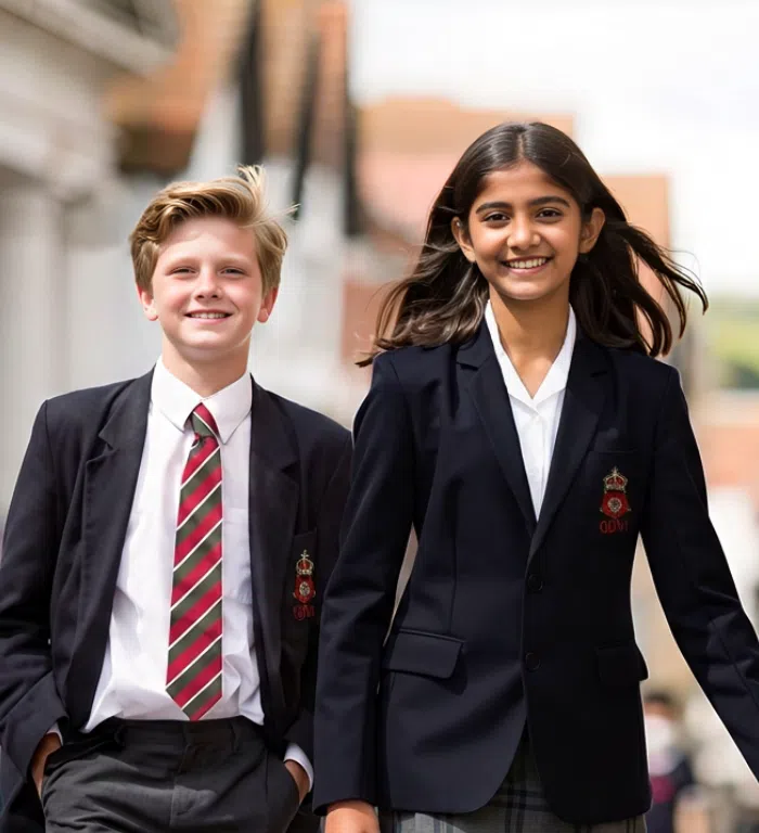 Two students in navy blazers walk outdoors on school grounds, smiling at the camera.