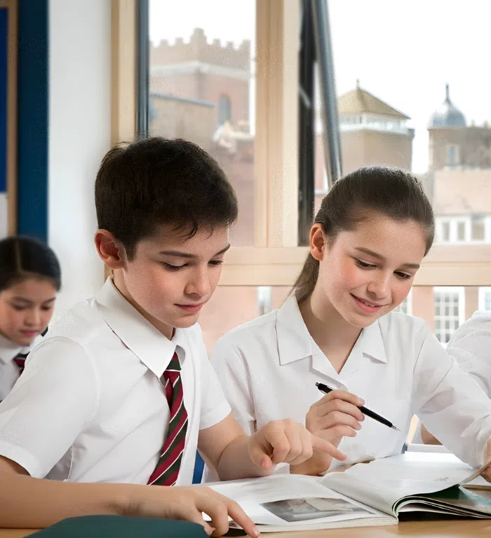 Two students in white shirts study together at a classroom desk, looking at an open textbook by a window.