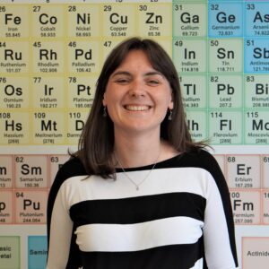Smiling woman standing in front of a colorful periodic table wall as a backdrop.