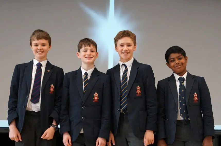 Four schoolboys in navy blazers and striped ties stand together, smiling for a photo in a classroom-like setting.