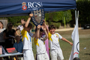 Children in white and yellow uniforms lift a shield trophy under a blue canopy.