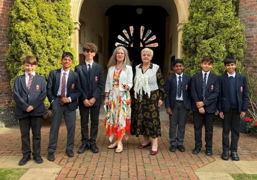 Group of students in navy blazers and ties flanked by two female teachers, posing outside a brick building with an arched doorway and greenery.