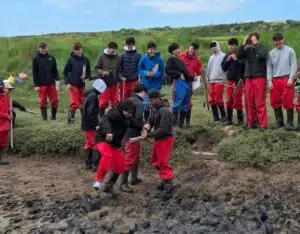 Group of students in red waterproof pants watch two peers digging in the muddy shoreline with small tools during an outdoor field activity.