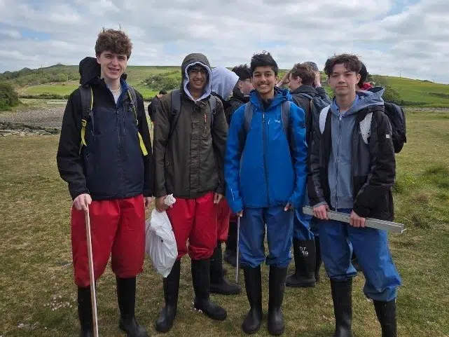 Five students in waterproof jackets and boots stand on a grassy field with rolling hills in the background, some carrying bags and a metal measuring tool as they pose for a field-trip photo.
