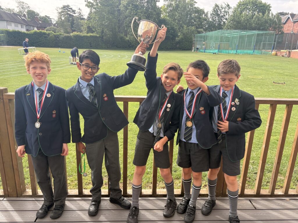 Five schoolboys in uniform lift a large trophy together on a wooden deck, smiling proudly.