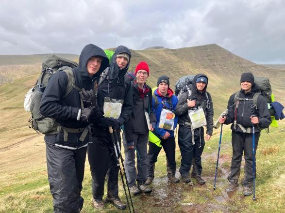 Six hikers in rain gear with large backpacks posing on a muddy trail in a grassy, hilly landscape; some hold maps.