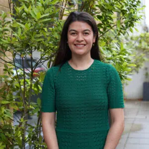 Smiling woman in a green textured dress standing outdoors beside a brick wall and green shrubs.
