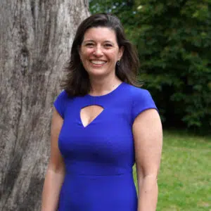 Smiling woman in a cobalt blue dress standing outdoors near a large tree and greenery behind her.