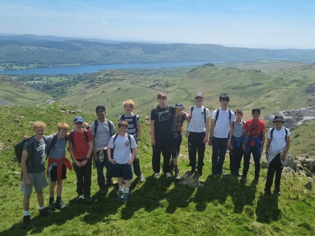 Group of students standing together on a grassy hilltop with backpacks, overlooking a green valley and a blue lake on a sunny day.