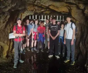 Group of teens with backpacks posing inside a rocky cave near a metal barrier.