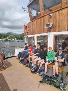 Group of kids sitting on the deck of a wooden boat by a lake, with backpacks on the ground nearby.