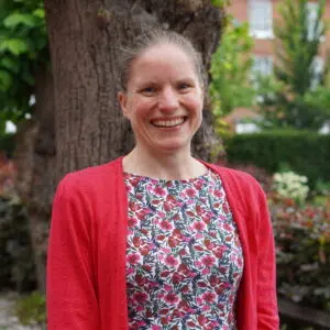 Smiling woman in a red cardigan and floral dress stands outdoors by a tree and garden.