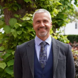 Smiling man in a navy vest and gray blazer stands outdoors with green leaves behind him.