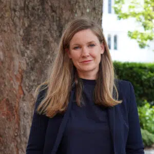 Portrait of a woman with long light brown hair standing outdoors beside a large tree, wearing a dark navy blazer and top.