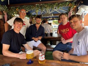 Five men sit at a wooden picnic table under a blue canopy in an outdoor pub, smiling with pints of beer in front of them.