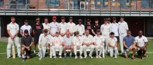 Group of cricket players posing for a team photo on a sunny grass field; many in white uniforms, some with bats and caps, arranged in two rows with seated players in front.