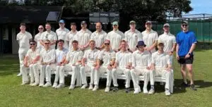 Group of cricket players in white uniforms posing for a team photo on a sunny field, with a scoreboard in the background.