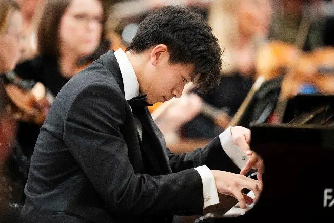 Male pianist in a black tuxedo playing a grand piano at a concert, with an attentive audience in the blurred background