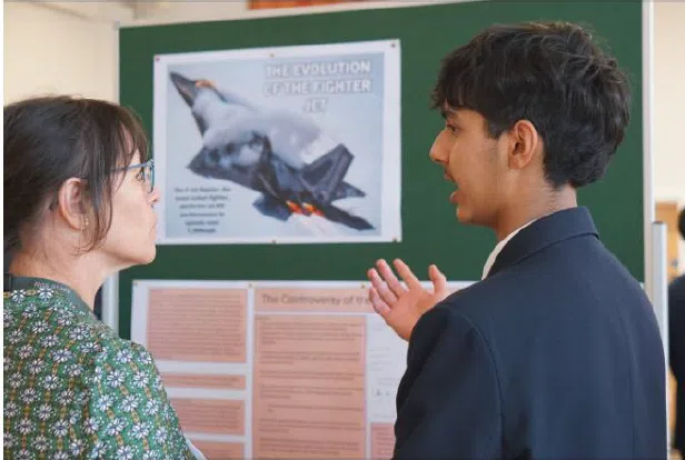 Two students discuss a science fair poster about the evolution of the fighter jet, displayed on a green board.