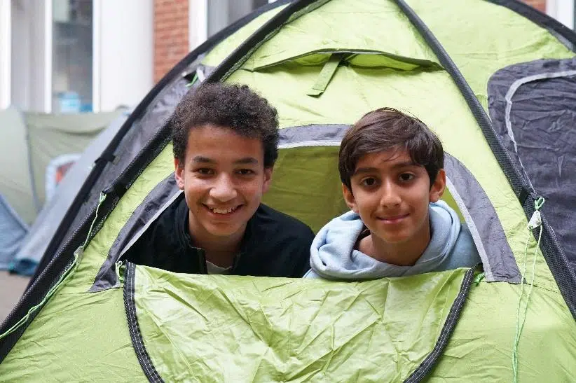 Two smiling boys lean out from a lime green tent, with other tents and buildings visible in the background.