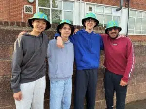 Four friends stand shoulder to shoulder outside a brick building, smiling and wearing matching blue patterned bucket hats and casual hoodies.