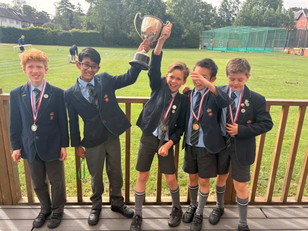 Five schoolboys in uniform on a deck hold up a large trophy and medals, celebrating a win.