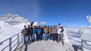 Group of diverse hikers posing together on a snowy mountain viewing deck, with a telescope and snow-covered peaks behind them under a blue sky.
