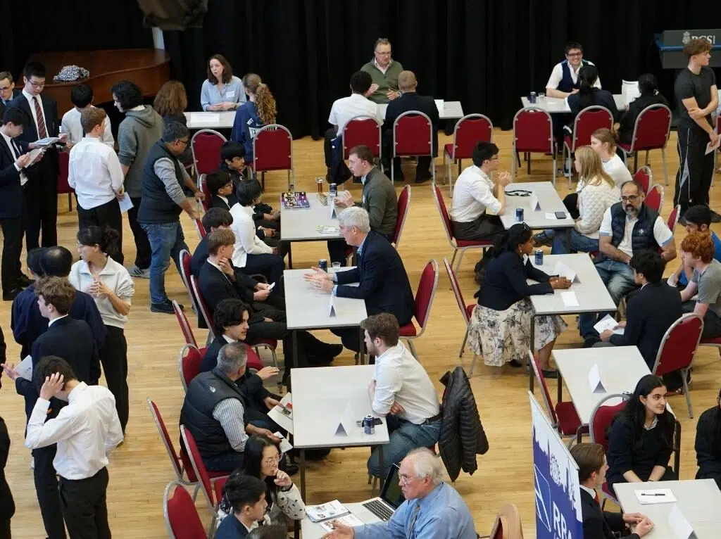 A crowded indoor event with many people seated at tables, likely a career fair or interview day, in a large hall with red chairs.