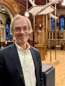 Smiling man in a navy blazer and white shirt standing in an ornately decorated church interior. (informative portrait)