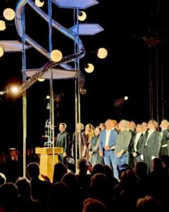 Nighttime formal event with a speaker at a wooden podium and a line of people in suits on stage, audience visible in foreground.