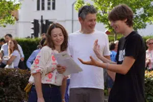 Three people conversing outdoors; a woman holds papers while two men smile and gesture as they talk.