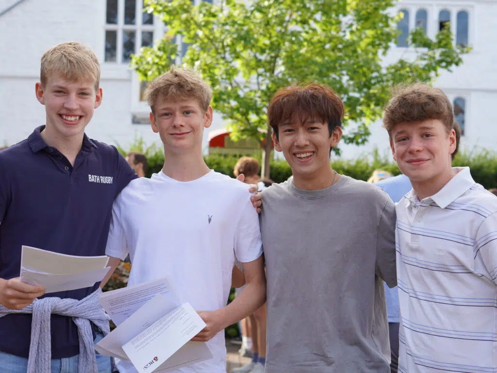 Four smiling young men stand together outdoors, holding papers, posing for a photo at an event.