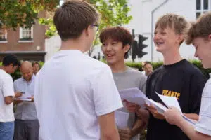 A group of young men talking and smiling outdoors, each holding papers, with a brick building and trees in the background.