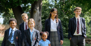 Group of smiling students in school uniforms posing outdoors under a leafy tree.