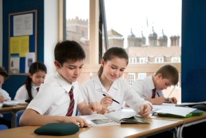 Students in white shirts and striped ties sit at desks, writing in notebooks in a bright classroom.